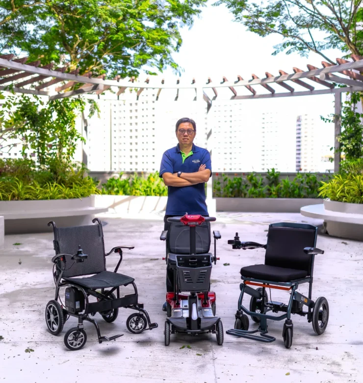 Warren Chew stands with three mobility devices: a power wheelchair, a red mobility scooter, and a manual wheelchair, in an outdoor setting.