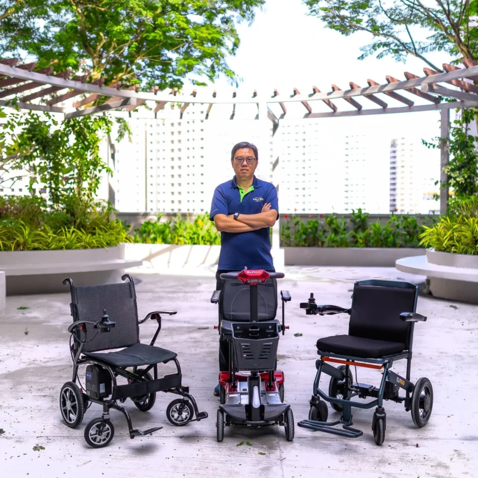 Warren Chew stands with three mobility devices: a power wheelchair, a red mobility scooter, and a manual wheelchair, in an outdoor setting.